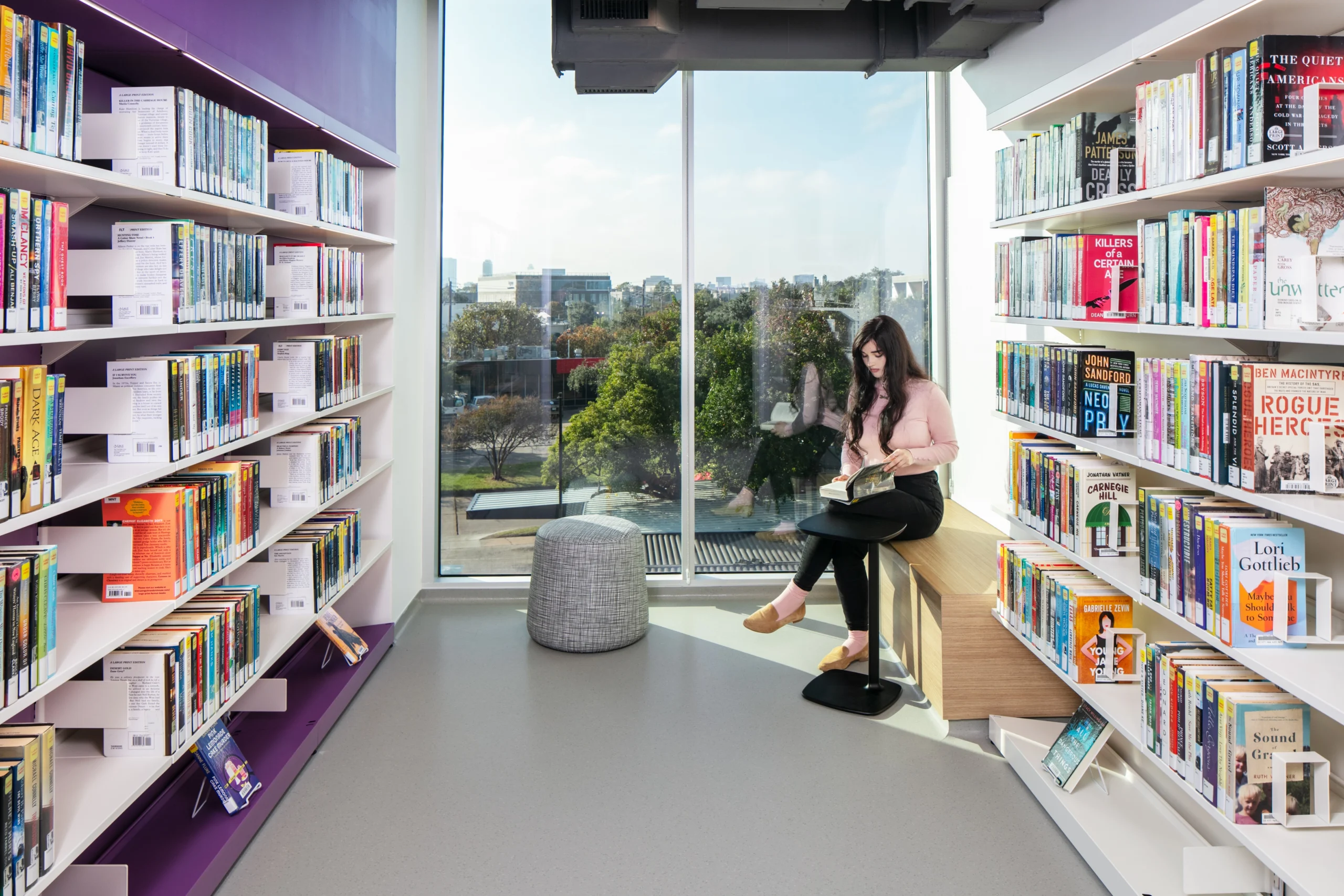 Girl sits in library with purple accent wall and floor to ceiling window looking to nature and skyline